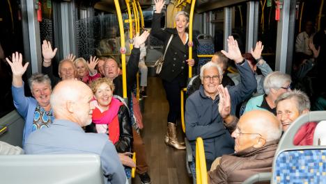 Personen sitzen und stehen im Bus und winken in die Kamera, gelbe Stangen und blau-graue Sitze, links und rechts Fensterscheiben, brauner Holzboden