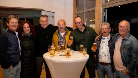 7 Personen stehen um Stehtisch mit weißer Husse, zwei halten Bier im Glas in der Hand, auf dem Stehtisch Deko und gefüllte Biergläser, Holzboden, im Hintergrund weiße Wand und rechts Fensterfront mit geöffneter Tür