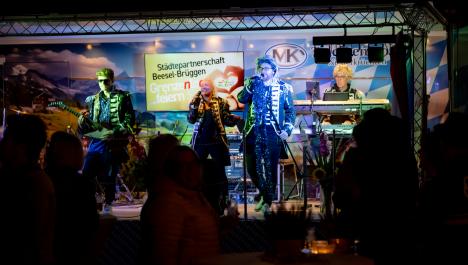 Band Schlagerzirkus auf Bühne, 5 Personen in Sakkos, teilweise mit Brille oder Perücke, rechts Mann am Keyboard, links Mann an Gitarre und Schlagzeug, mittig Frau und Mann mit Mikrofonen, Bühne bayrisch geschmückt, Rückwand mit weiß-blau karierter Abdeckung, Werbung für Trachtenmode Niederkrüchten, links Bildschirm mit „Städtepartnerschaft Beesel-Brüggen Grenzenloos feiern“, vorne Schatten von Menschen