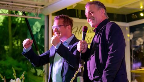  Bürgermeister Bob Vostermans (Beesel, links) mit Brille hellblauen Hemd, blauen Sakko hält gedrückte Daumen nach vorne, Bürgermeister Frank Gellen (Brüggen, rechts) in schwarzen Hemd und Sakko hält Mikrofon vor sich in rechter Hand, m Hintergrund dekorierte Bühne