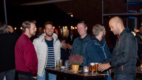 5 Männer stehen an braun-schwarzen Stehtisch, mittig lila Blume in braunen Topf und Teelicht in Glas, auf Tisch gefüllte Biergläser, im Hintergrund Schatten von Menschen und beleuchtetes Gebäude