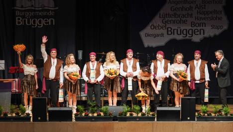 Menschengruppe in Piratenkostümen steht auf einer Bühne und schauen in die Kamera. Im Hintergrund sieht man ein dunkles Plakat mit der Logo der Burggemeinde Brüggen und einem Kartenumriss mitsamt der Schriftzüge der Ortsnamen der Gemeinde. Rechts steht der Bürgermeister und applaudiert. Im Vordergrund sind Tänzerinnen und Tänzer mit brauner und weißer Kleidung. Die Herren tragen rote Kopfbänder und die Damen halten einen orangen Blumenstrauß in der Hand. Links steht ein Herr, der in die Kamera winkt.