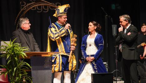 Das Brüggener Prinzenpaar steht auf der Bühne mit dem Bürgermeister rechts und einem weiteren Herr links im Bild. Der Herr links und der Bürgermeister tragen beide einen schwarzen Anzug und schauen zu dem Prinzenpaar. Der Prinz trägt eine weiße Hose und eine blaue Kutte mit goldenen Verzierungen. Sein Hut ist ebenfalls gold und blau und hat lange goldene Federn, die nach hinten ausgerichtet sind. Die Prinzessin trägt ein blau weißes Kleid und hat die Haare nach hinten gesteckt.