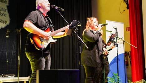 Ein Mann und eine Frau stehen auf der Bühne und musizieren. Links im Bild steht der schwarz gekleidete Herr, hält seine Gitarre und singt ins Mikrofon. Rechts neben ihm steht eine Frau ebenfalls in schwarzer Kleidung und hält das Mikrofon in der Hand und singt.