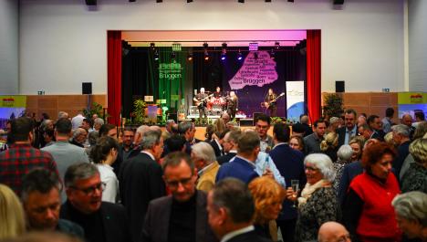 Eine Menschenmenge ist versammelt vor einer grün und pink leuchtenden Bühne. Auf der Bühne stehen 2 schwarz gekleidete musizierende Menschen mit Gitarren, Schlagzeug und Mikrofonen. Im Hintergrund der Bühne sieht man das Logo und den Kartenumriss der Burggemeinde Brüggen.