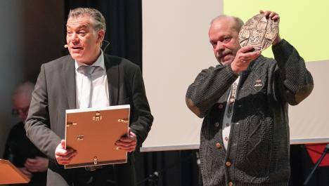 Der Bürgermeister Frank Gellen hält links im Bild eine Urkunde in der Hand. Er trägt einen schwarzen Anzug und ein weißes Hemd mit Krawatte. Rechts von ihm hält der ein Herr mit einer grauen Strickjacke die Ehrenplakette der Burggemeinde in die Luft und lächelt. Im Hintergrund sieht man eine Leinwand.