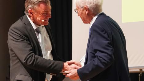 Der Bürgermeister Frank Gellen steht mit einem schwarzen Anzug links im Bild. Er hat ein Mikrofon an. Rechts im Bild ist ein Mann mit Brille im blauen Anzug zu sehen, der eine Urkunde in der Hand hält. Beide geben sich die Hand und schauen sich an.
