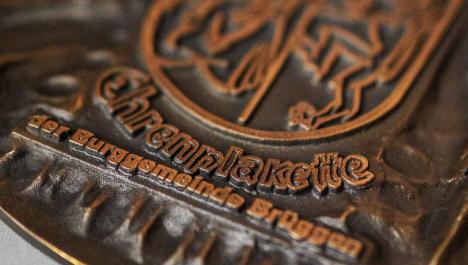Eine Nahaufnahme der bronzefarbenen Ehrenplakette der Burggemeinde Brüggen. Der Schriftzug der Ehrenplakette ist unterhalb des Brüggener Wappens gedruckt. Die Plakette ist rund.