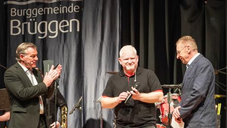 Der Bürgermeister steht links im Bild und applaudiert während mittig ein Sänger steht, der ein Mikrofon in der Hand hält. Recht steht ein weiterer Herr mit einem Umschlag in der Hand. Alle drei Herren sind dunkel gekleidet und stehen auf der Bühne vor Blas- und Schlaginstrumenten. Im Hintergrund ist ein blauer Vorhang mit dem Logo der Burggemeinde Brüggen.