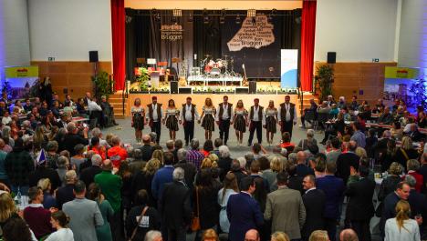 Eine Gruppe von Tänzerinnen und Tänzern steht aufgereiht nebeneinander vor einer Bühne und halten sich bei den Händen. In einem Halbkreis stehen Menschen um die Gruppe herum und applaudieren. Die Tanzgruppe trägt Piratenkostüme in braun und weiß, sowie rote Kopfbänder. Im Hintergrund sieht man die Bühne mit Instrumenten und einem Vorhang mit Logo der Burggemeinde Brüggen.