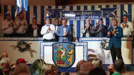 Die Brachter Wasserratten und der Bürgermeister Frank Gellen stehen vorne auf der Bühne und gucken in die Kamera. Außerdem klatschen die Leute auf der Bühne. Im Hintergrund ist eine blau und weiß gestreifte Wand. Im Vordergrund ist ein großen Wappen zu sehen.