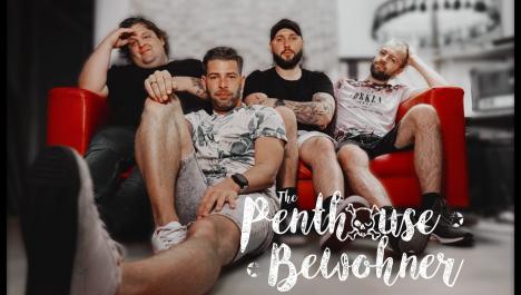 Auf einer roten Couch sitzen vier Männer in einem Raum mit hellen weißen Wänden. Rechts unten im Bild ist Schriftzug der Band eingeblendet. Links auf der Couch sitzt ein Mann mit schwarzem T-Shirt und dunkler Jeans. Rechts neben ihm sitzt ein Mann mit weißem T-Shirt und einer kurzen grauen Hose. In der Mitte sitzt ein Herr mit schwarzer Kappe und T-Shirt und einer hellen kurzen Hose. Rechts auf der Couch sitzt ein Herr mit Brille und einem weißem T-Shirt mit Aufdruck und einer schwarzen kurzen Hose.