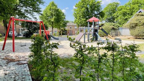 Ein Spielplatz ist zu sehen. Links im Bild steht eine rote Schaukel und rechts im Bild ein hellblaues Klettergerüst mit einer schwarzen Rutsche. Im Vordergrund ist eine grüne Hecke zu sehen. Im Hintergrund stehen Wohnhäuser, eine Kletterwand sowie zahlreiche grüne Bäume. Der Untergrund besteht aus Sand. Oben im Bild ist ein blauer Himmel mit einzelnen Wolken.