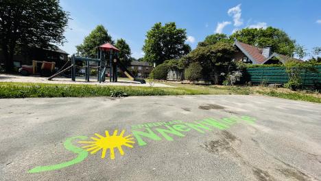 Auf einer asphaltierten Fläche ist ein grüner Schriftzug Spielplatz Sonnenwiese aufgedruckt mit einer gelben Sonne, die das O ersetzt. Im Hintergrund ist das hellblaue Klettergerüst mit rotem Dach abgebildet, sowie steht rechts ein Haus. Ebenfalls im Hintergrund ist ein grüner Rasen, grüne Bäume und ein blauer Himmel mit vereinzelten kleinen Wolken zu sehen.