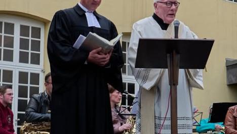 Zwei Pfarrer, links in schwarz gekleidet und rechts in weiß gekleidet, halten eine Ansprache auf einer Bühne. Im Hintergrund sieht man eine Musikgruppe und das gelbe Rathaus der Burggemeinde Brüggen.