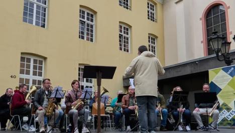 Eine Musikgruppe gibt ein Konzert auf einer Bühne. Im Vordergrund stehen Notenständer und der Dirigent. Im Hintergrund sieht man das gelbe Gebäude des Rathauses in Brüggen. Rechts im Hintergrund sieht man ein Stück des Kirchengebäudes des St. Nikolaus in Brüggen.