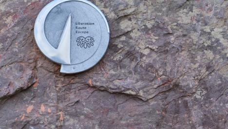 Ein Hörstein ist an einer braunen Steinwand festgehakt. Auf dem silbernen Siegel des Steines steht Liberation Route Europe.