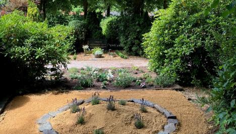 Das Sandarium ist zu sehen. Hierbei handelt es sich um einen Steinkreis auf einem Sandhügel für Insekten, die im Boden leben. Im Steinkreis sind ein paar Pflanzen eingesetzt worden. Im Hintergrund wurde ein Grünstreifen ebenfalls mit Blumen bepflanzt. Im Hintergrund sind Bäume und Hecken zu sehen.