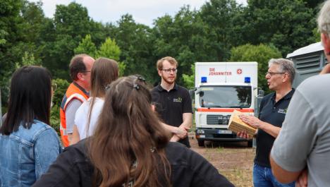 Ein weißer LKW der Johanniter ist im Hintergrund vor grünen Bäumen platziert. Im Vordergrund stehen zahlreiche Menschen.