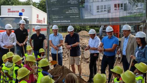 Architekten, Vertreter/innen der Johanniter und der Gemeinde Brüggen haben weiße Helme auf und stehen vor einer Absperrung. Im Vordergrund sind Kinder in gelben Helmen zu sehen, die mithilfe von Spaten in die Erde stechen.