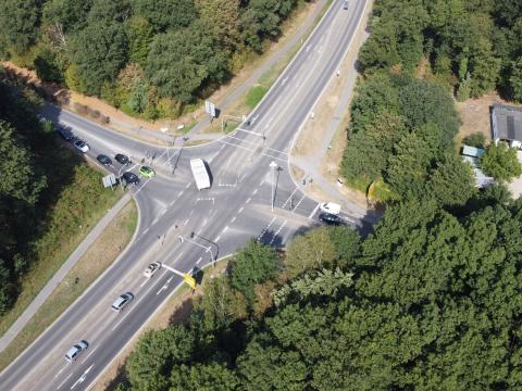 Luftbild von ursprünglicher Ampelkreuzung B221/Borner Straße/Boisheimer Straße mit weißen Fahrbahnmarkierungen, Fußgängerwegen und Grünstreifen am Straßenrand umgeben von dichtem, verschiedenartigen Baumbewuchs vereinzelt Autos auf der Straße, oben rechts zwischen den Bäumen Wohnhaus mit Grundstück