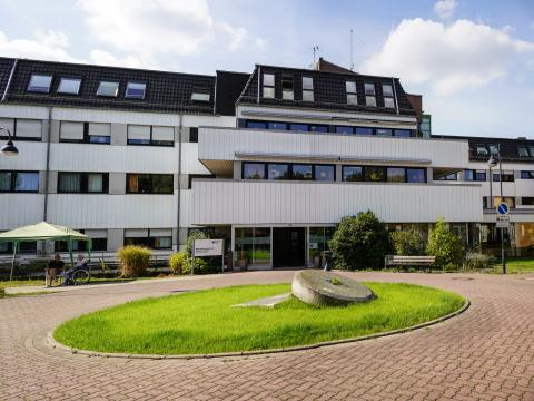 Frontansicht auf weißes Gebäude des DRK Seniorenzentrum Brüggen-Bracht mit Balkonen und Fenstern, vor dem Gebäude gepflasterter Weg mit einer runden Grasfläche und großen, runden Stein in der Mitte, vor Gebäude Wieseflächen und vereinzelter Strauchbewuchs, rechts eine Sitzbank, links sitzen zwei Personen, eine im Rollstuhl, unter einem Pavillon