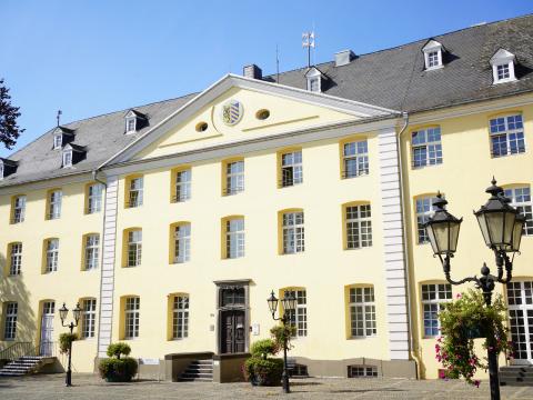 Gelbes historisches Gebäude "Rathaus Brüggen" mit Treppenaufgang zur braunen Eingangstür am gepflasterten Kreuzherrenplatz, Ausschnitt von Baumkrone, Schatten des Baumes auf Platz, auf dem Platz vereinzelt Straßenlaternen mit Hängepflanzen und Sitzbänke