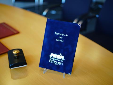 Blaues Stammbuch mit weißer Aufschrift "Stammbuch der Familie" und weißes Logo der Burggemeinde Brüggen stehend in einer Halterung auf einem Tisch, links davon schwarz, weiße Schatulle mit goldenen Pin oben drauf, im Hintergrund in der oberen, rechten Ecke Stühle am Tisch 