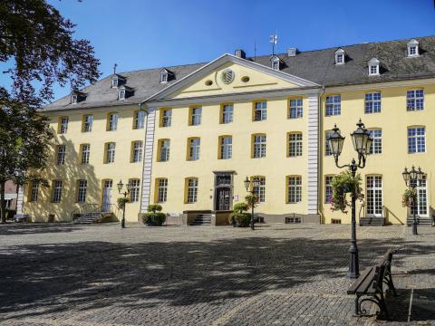 Gelbes historisches Gebäude Rathaus Brüggen mit Treppenaufgang zur braunen Eingangstür am gepflasterten Kreuzherrenplatz, Ausschnitt von Baumkrone, Schatten des Baumes auf Platz, auf dem Platz vereinzelt Straßenlaternen mit Hängepflanzen und Sitzbänke