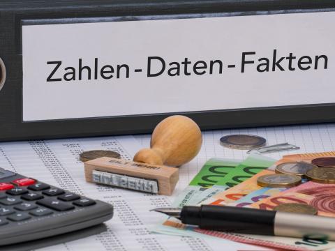 Schwarzer, breiter Aktenordner beschriftet mit "Zahlen-Daten-Fakten", davor liegen Taschenrechner, Geld in Münzen und Scheinen, Stempel auf Blättern, teils beschriftet