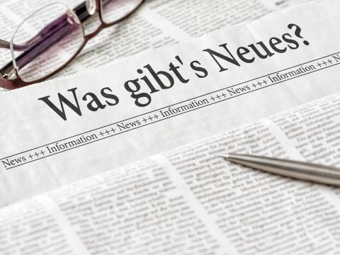 Beschriebene Zeitungsseiten übereinander gestapelt, in der Mitte Zeitungsseite mit der Überschrift: Was gibt´s Neues? Auf den Zeitungspapieren liegt oben eine Brille und unten rechts ein Kugelschreiber. 