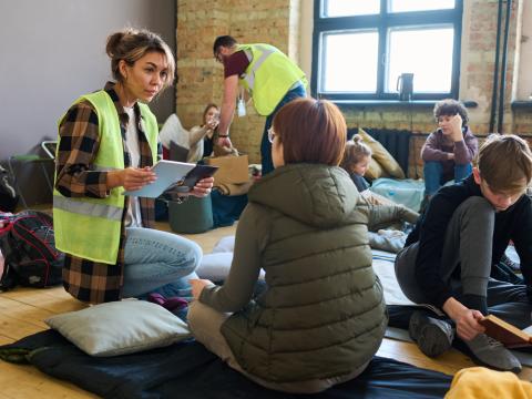 Freiwillige auf Knien mit gelber Warnweste und mobilen Geräten kommuniziert mit einem der Flüchtlinge auf dessen Schlafplatz in Unterkunft für Migranten, im Hintergrund weitere Schlafplätze, Taschen, ein Helfer und Flüchtlinge 