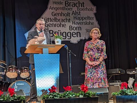 Bürgermeister Frank Gellen am Rednerpult mit Mikrofon, rechts davon Frau mit bunten Blumenkleid auf Bühne mit rotblühenden Blumen in Blumenkästen vorne, Schlagzeug, Mikrofone und Vorhang dahinter