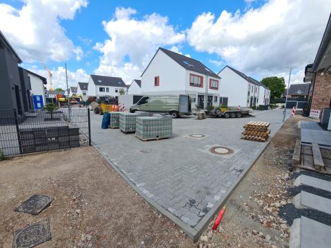 Wohngebiet mit neugebauten (Doppel-) Häusern und gepflasterter Straße, worauf Palletten mit Steinen stehen, rechter Auszug vom Bild zeigt Neubau mit unfertigen Vorgarten, linker Auszug vom Bild zeigt erdige Freifläche an einem Zaun bei leichtbewölktem, blauen Himmel 