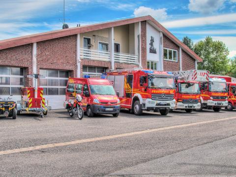 Feuerwehrgerätehaus Brüggen: Gebäude mit Balkon und geschlossenen Garagen, davor auf großer Einfahrt (von links nach rechts) Quad, Anhänger mit Lichtmast, Motorrad, Mannschaftstransportfahrzeug, Löschgruppenfahrzeug, Drehleiterfahrzeug, Löschgruppenfahrzeug und Mehrzweckfahrzeug in rot bei blauem, leicht bewölktem Himmel