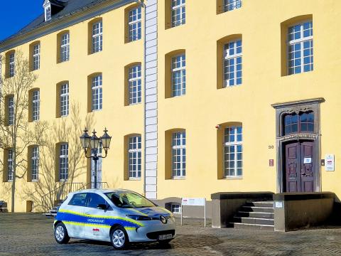 Weißer Elektro-Kleinwagen mit Schriftzug "Ordnungsamt" auf blauem Streifen gelb umrahmt, darunter Logo der Burggemeinde Brüggen mit Treppenzugang vor gelben Gebäude auf gepflasterten Platz 