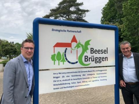 Schild zwischen blauen Rahmen mit Überschrift in blau "Strategische Partnerschaft", darunter Logo Beesel-Brüggen (Umriss Burg orange, unter linker Teil der Burg drei grüne Bäume, rechts davon blauer Halbkreis mit gelben Sternen als Zeichen der Europäischen Union, rechts von der Burg grüner Drache, rechts Schriftzug "Beesel Brüggen") und Informationstext darunter, links und rechts davon je ein Mann im Anzug , im Hintergrund gepflasterter Platz und dichter, grüner Baumbewuchs 