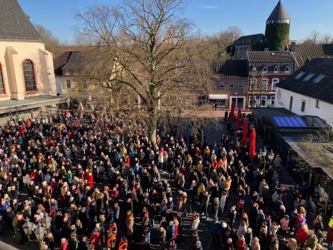 Bunte Menschenmenge teils mit Schildern auf Kreuzherrenplatz vorm Rathaus, mittig großer kahler Baum, links Ausschnitt von Katholische Kirche St. Nikolaus, rechts Gebäude mit roten Schirmen und Sitzgarnituren draußen, im Hintergrund weitere Gebäude, Fußgängerzone, Burg Brüggen, kahle Bäume und blauer Himmel