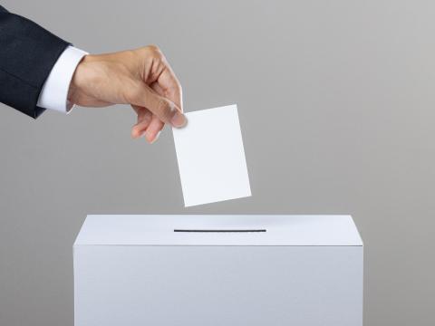 Links im Bild hält jemand einen weißen Wahlschein in der Hand und wirft diesen in eine hellgraue Wahlurne ein. Ebenfalls links im Bild sieht man ein Stück von dem Ärmel der Person, die ein weißes Hemd unter einem schwarzem Anzug trägt. Der Hintergrund ist ein grauer Farbverlauf.