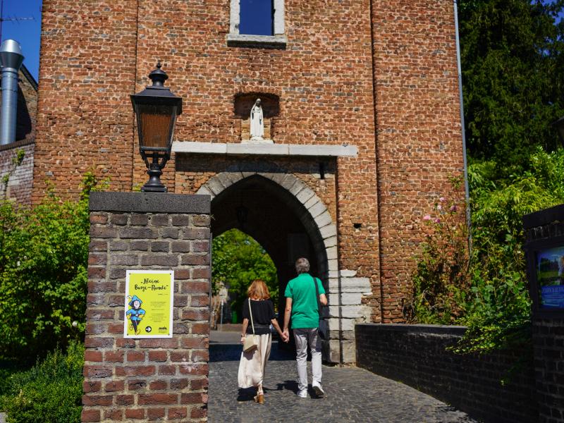 Zwei Spaziergänger auf einer gepflasterten Brücke vor dem Tor zu Burg Brüggen, vor der Brücke rechts und links zwei gemauerte, rechteckige Pfeiler mit Laterne oben drauf, seitlich Plakate und dahinter dichtem, grünen Strauchbewuchs