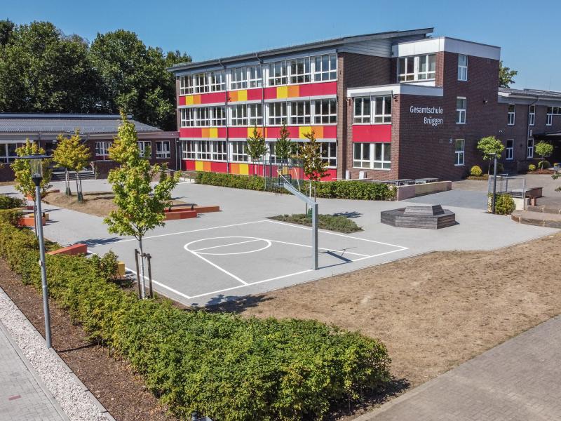 Schulhof mit Sitzgelegenheiten, Bäumen und weißen Linien für Basketballfeld mit Wurfkorb vor dem Gebäude der Gesamtschule Brüggen, mit weißen Fensterrahmen und rechts weißen Schriftzug "Gesamtschule Brüggen" und rot, orangen und gelben Platten zwischen Fenstern links, im Vordergrund grüne Hecke und sandiger Boden, links Ausschnitt von Parkplatz, im Hintergrund dichter, grüner Baumbewuchs, blauer Himmel