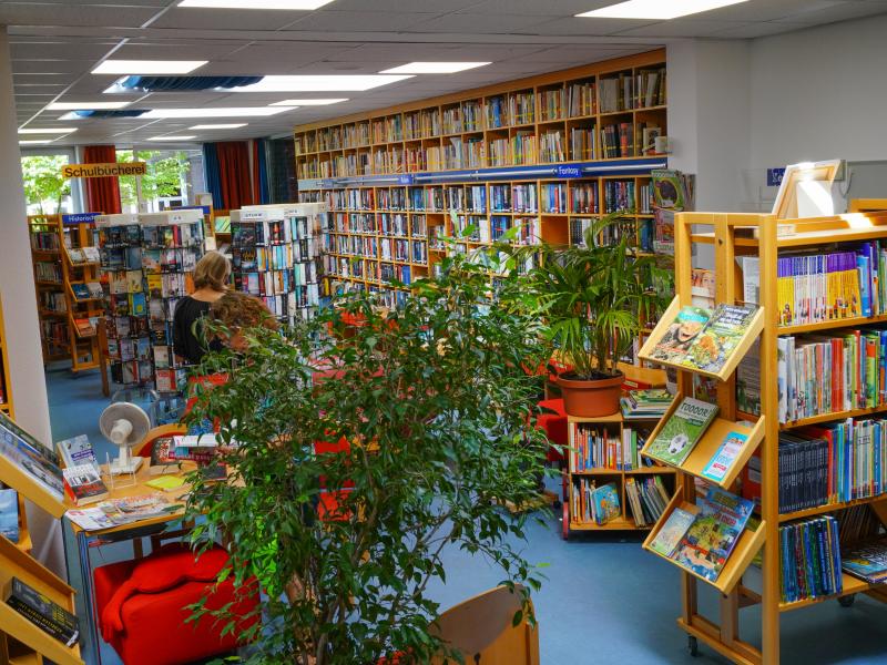 Person in Bücherei zwischen vielen hölzernen Bücherregalen, -aufstellern und Pflanzen mit verschiedenen, bunten Büchern, blauer Boden und weißer Decke mit leuchtenden Lampen