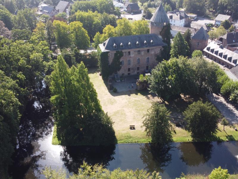 Panoramabild von Burg Brüggen mit teils trockener Burgwiese, Burgweiher und dichtem, teils vereinzelten Baumbewuchs, rechte obere Ecke mehrere Gebäude