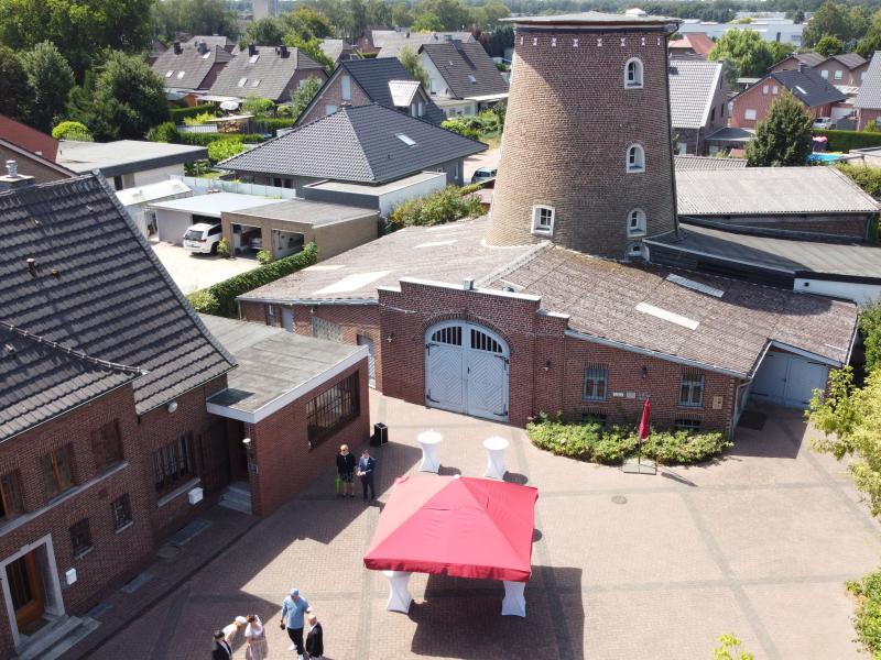 Panoramablick auf die Brachter Mühle: im Vordergrund links Gebäude, rechts davor gepflasterter Platz mit rotem Pavillon, Stehtischen mit weißen Hussen und Menschen, rechts grüner, dichter Baumbewuchs, mittig Gebäude der Brachter Mühle mit weißem Tor, im Hintergrund weitere Gebäude und vereinzelter Bäumen