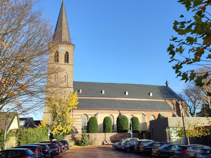 Parkplatz mit geparkten Autos links und rechts, vereinzelter Baum- und Heckenbewuchs vor einer Kirche mit Kirchturm auf der linken Seite