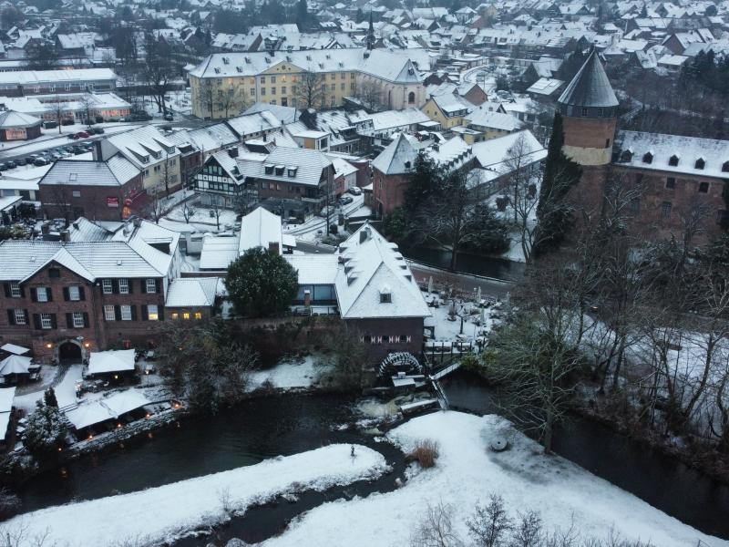 Panoramablick über den schneebedeckten Ortseingang und Ortskern von Brüggen im Winter: vorne links Gebäude der Torschänke mit Tischen, Stühlen und Schirmen davor, daneben rechts Gebäude Brüggener Mühle, vor den Gebäuden Fluss "Schwalm" mit schneebedeckter Wiese, kahlem Strauch- und Baumbewuchs, rechts Burg Brüggen davor kahler Baumbewuchs, im Hintergrund weitere schneebedeckte Gebäude