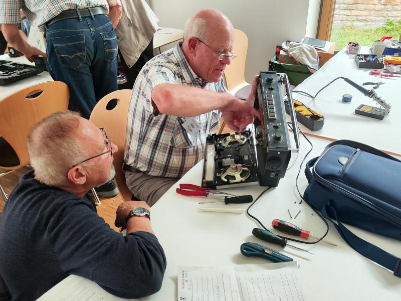 Mittig: Älterer Mann mit Brille und kariertem Hemd sitzt an weißem Holztisch und schraubt an elektronischen Gerät, rechts von ihm Mann mit Brille, Armbanduhr, und Pullover schaut zu, auf Tisch diverse Werkzeuge und ein blauer Rucksack, im Hintergrund links stehen und sitzen Personen an weißem Holztisch und auf Holzstühlen mit Metallbeinen und rechts Holzkiste und Werkzeug auf Tisch vor Fenster mit Holzrahmen mit Blick auf Bachsteinwand und Wiese