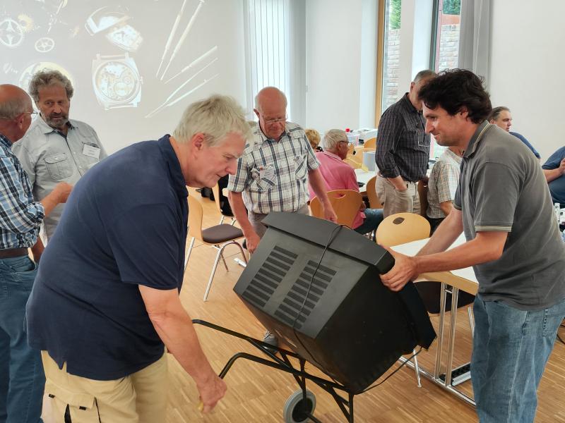 Im Vordergrund: Mann links graue Haare, blaues Polohemd, beige Hose trägt mit Mann rechts, dunkle Haare, graues Polohemd und Jeans einen Kastenfernseher auf Gestell mit Rollen, im Hintergrund links unterhalten sich zwei Männer in Hemden, dahinter auf Leinwand eine auseinander gebaute Uhr mit Werkzeug projiziert, rechts und mittig sitzen auf Stühlen und stehen Männer und Frauen an weißen Holtischen mit verschiedenen Utensilien und Werkzeuge, dahinter weiße Wand, zwei Fenster mit Blick auf Backsteinwand 
