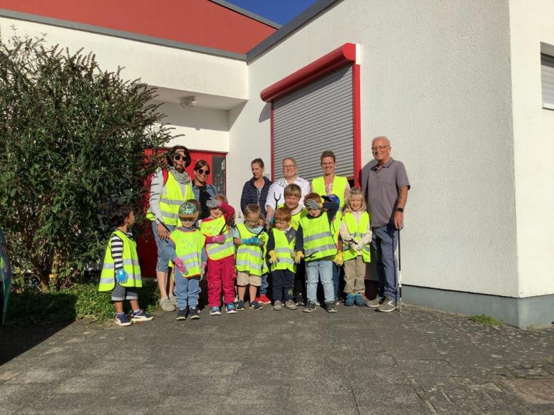 Gruppe von Kindern mit gelben Warnwesten und 6 Erwachsenen, teils mit Warnwesten, stehen vor rot-weißem Gebäude, hinter der Gruppe rote Eingangstür, rechts davon Fenster mit Rollos halb unten und roten Fensterrahmen, links grüner Busch, vor dem Gebäude Steinplatten