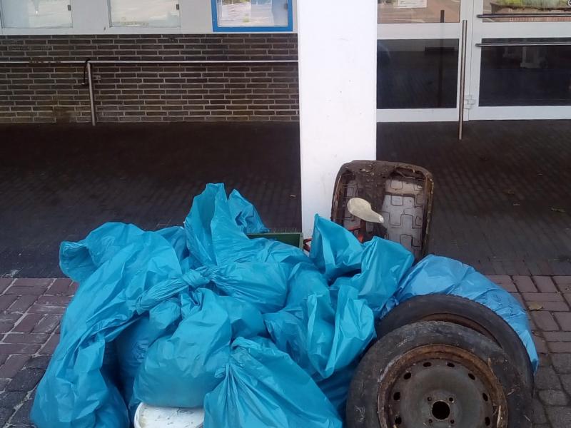 Müllberg mit blauen Müllsäcken, Autoreifen und weißem Farbeimer auf gepflastertem Boden, vor weißer Säule mit Logo vom World Cleanup Day und Burggemeinde Brüggen, im Hintergrund links drei Glasschaukästen mittig an der Wand und rechts Glastür mit weißem Rahmen